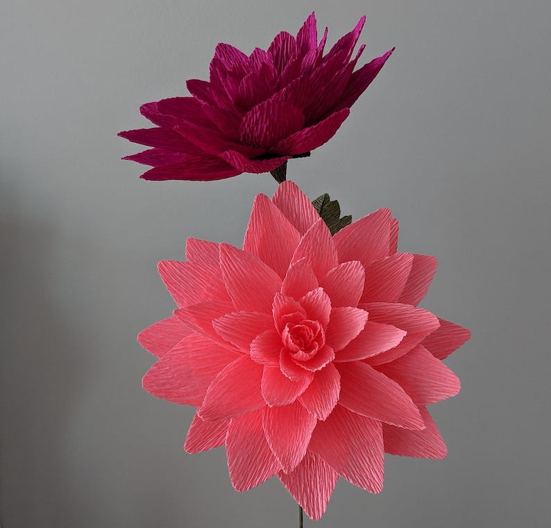 May include: Two large paper flowers, one pink and one magenta, are arranged together on a stem. The flowers are made of crinkled paper and have a layered, ruffled appearance.