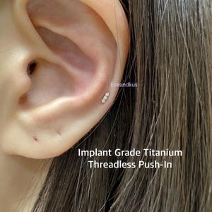 May include: An ear with a small, curved bar earring featuring three clear, round gemstones. The earring is gold-coloured. Text below the ear reads "Implant Grade Titanium Threadless Push-In".