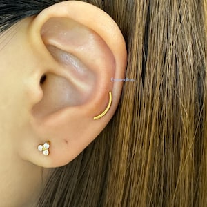 May include: A close-up of a person's ear with two gold earrings. The earring in the upper ear is a curved bar, and the earring in the lower ear is a cluster of three small, clear stones.