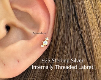 May include: Close-up of a person's ear with a small, internally threaded labret earring. The earring is made of 925 sterling silver and features three small, clear gemstones.