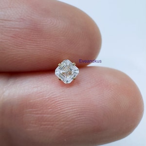 May include: A small, square-cut clear gemstone set in a gold-toned four-prong setting. The stone features a faceted, step-cut design that creates a geometric pattern. The text 'Eveandkus' is printed in small blue letters next to the gemstone.