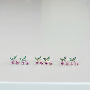 May include: Three pairs of earrings featuring a cherry design with green leaves and pink gemstones.