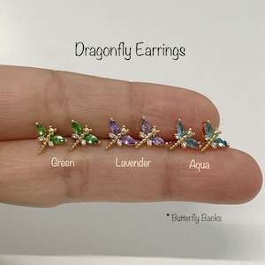 May include: Three pairs of dragonfly earrings with butterfly backs. The earrings are gold-toned and feature a dragonfly design with green, lavender, and aqua colored gemstones.