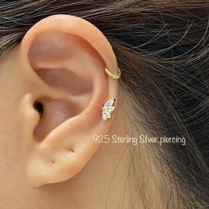 May include: A close-up of a person's ear with a small, gold, cz-studded earring in the helix piercing. The earring is shaped like a cluster of small, marquise-shaped stones. The person also has a gold hoop earring in the upper lobe. The text "925 Sterling Silver piercing" is visible below the ear.