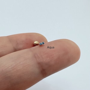 May include: A gold body piercing with two small blue gemstones. The piercing is labeled "Aqua".