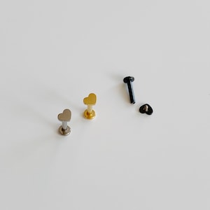 May include: Three heart-shaped body jewelry pieces. One is gold, one is silver, and one is black.