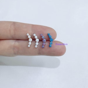 May include: Four sets of ear climbers with a curved design. Each set features multiple round, iridescent stones in white, purple, and blue. The earrings are displayed on a light background.