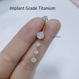 May include: A gold-colored belly button ring with a curved barbell design. The ring features a large, clear, round-cut stone at the top and a dangling chain of three smaller stones. The text "Implant Grade Titanium" is visible.