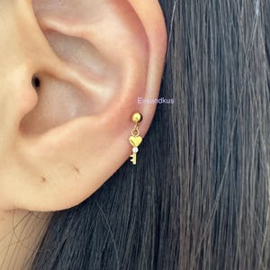May include: A gold-tone stud earring featuring a small heart-shaped charm attached to a dangling key pendant with a tiny clear stone accent. The earring is positioned in an earlobe, and the text Eveandkus is visible near the jewelry piece.