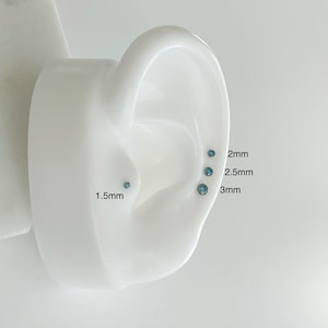 May include: A white ear model with five blue gemstone studs of different sizes. The sizes are labeled 1.5mm, 2mm, 2.5mm, and 3mm.