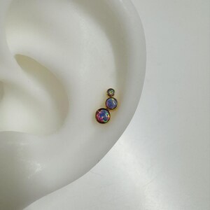 May include: Three gold ear studs with iridescent opal stones. The stones are different sizes and are arranged in a staggered pattern.