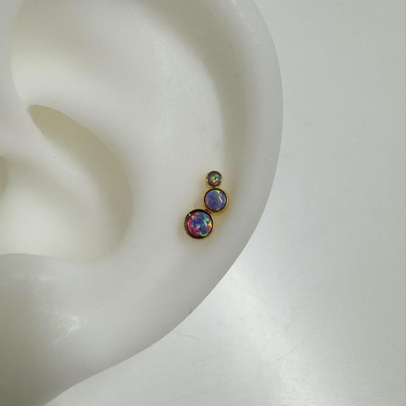 May include: Three gold ear studs with iridescent opal stones. The stones are different sizes and are arranged in a staggered pattern.