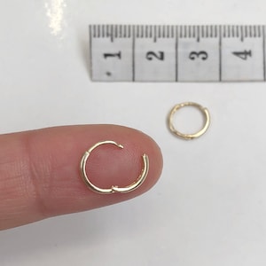 May include: Two gold hoop earrings, one is being held by a finger, the other is on a white surface. The earrings are approximately 1 inch in diameter.