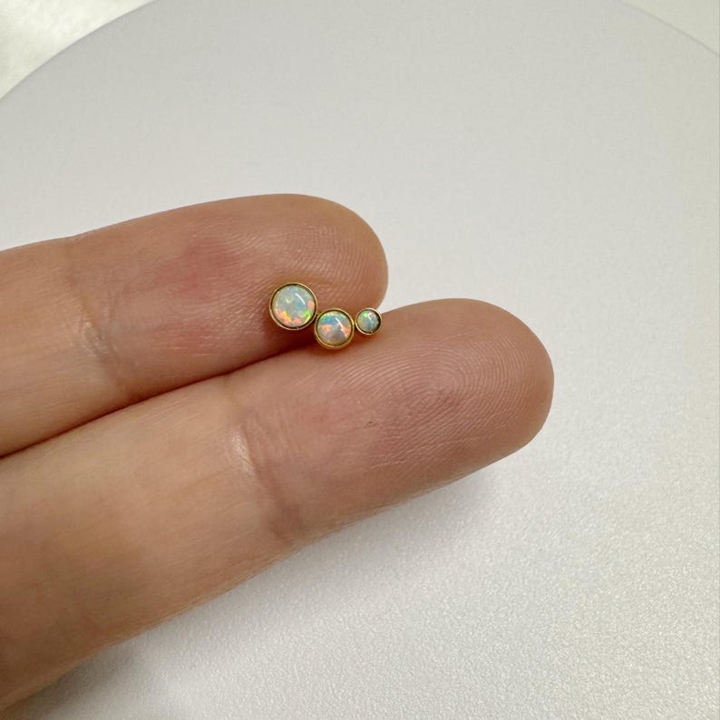 May include: Three gold-toned stud earrings with white opal stones. The stones are different sizes, with the largest stone in the center.