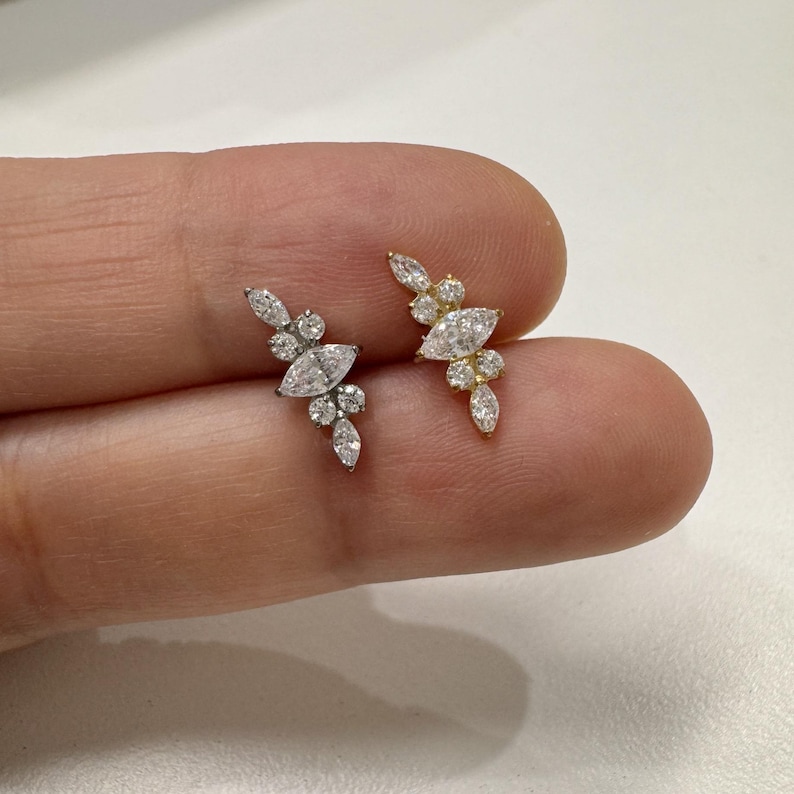 May include: Two delicate earrings, one silver and one gold, each adorned with a cluster of sparkling, clear gemstones. The design features a central marquise-cut stone surrounded by smaller, round and marquise-cut gems, creating a floral or leaf-like pattern.