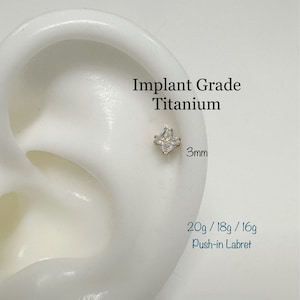 May include: A close-up of a clear cubic zirconia labret stud with a gold base. The stud is 3mm in size and is made of implant grade titanium. The text "Implant Grade Titanium" and "20g / 18g / 16g Push-in Labret" is visible in the image.