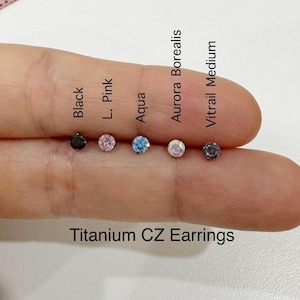 May include: Close-up of six titanium CZ earrings with different colored stones. The stones are black, light pink, aqua, aurora borealis, and vitrail medium.
