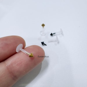 May include: A selection of lip piercing studs. One stud is in focus, with a clear post and a gold-coloured arrowhead. Other studs have black or gold arrowheads. The studs are on a white background.