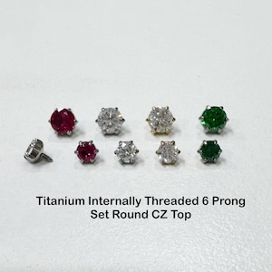 Può includere: Titanium internally threaded 6 prong set round CZ top. Una collezione di piccoli gioielli circolari con sei griffe metalliche che trattengono pietre rotonde nei colori rosso, trasparente e verde, disposti in due file su una superficie bianca.