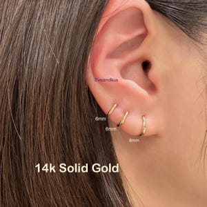 May include: Three gold hoop earrings, 6mm and 8mm in size, are shown on an ear. The earrings are labeled as 14k solid gold. The brand name "Eveandkus" is visible in the image.