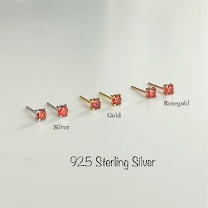 May include: Three pairs of small stud earrings with red gemstones. The earrings are shown in silver, gold, and rose gold.
