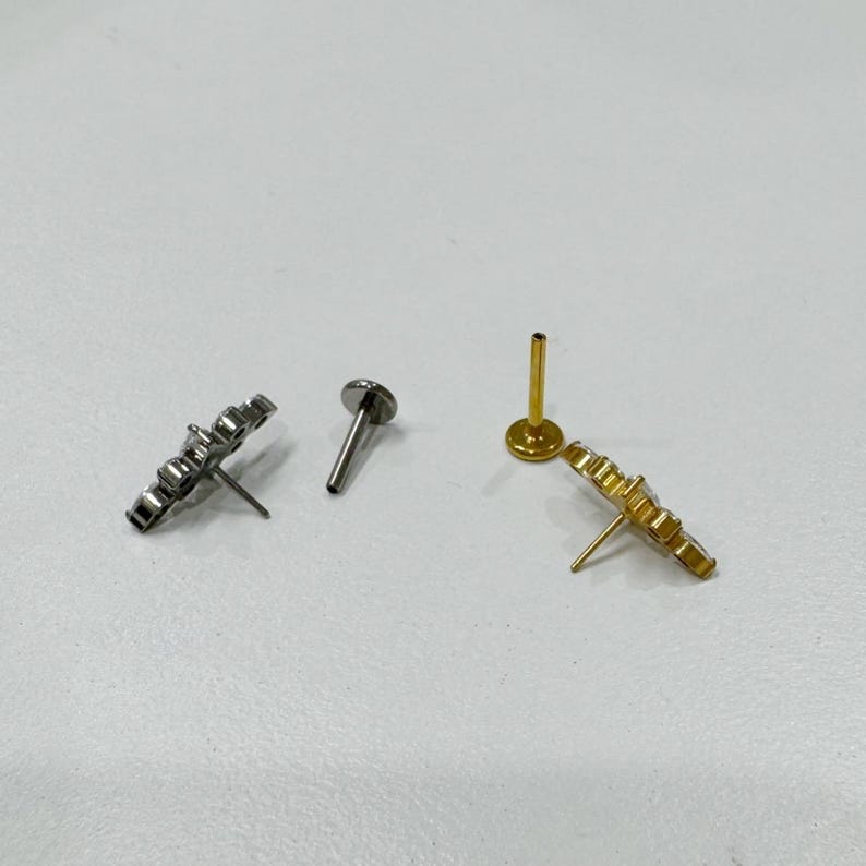 May include: Two stud earrings, one silver and one gold, each with a flower-like design. The silver earring has a flat back and a post. The gold earring has a flat back and a post. The earrings are on a white surface.