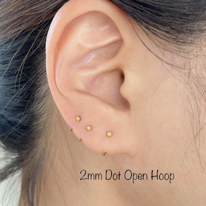 May include: An ear with several small, gold-coloured stud earrings and a tiny open hoop earring. The image includes the text "2mm Dot Open Hoop", indicating the product's style and size. The studs are approximately 2 millimeters in diameter.