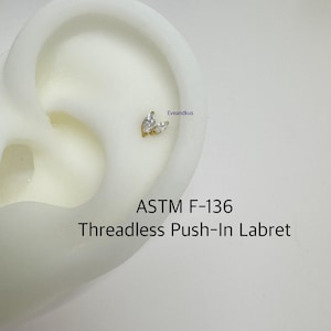 May include: A gold-colored threadless push-in labret stud with two clear, marquise-cut gemstones. The labret is shown on an ear. The text "ASTM F-136" and "Threadless Push-In Labret" are visible.