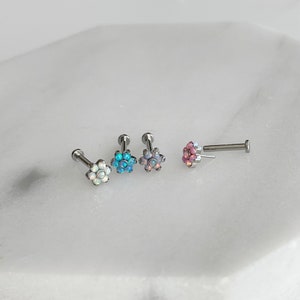 May include: Four flower-shaped body jewellery pieces in different colours. The jewellery is made of stainless steel and has a small flower design with opal-like stones. The colours are white, blue, pink, and a combination of pink and white.
