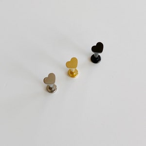 May include: Three heart-shaped lip studs in silver, gold, and black. The studs have a flat back and a heart-shaped top. The silver stud is on the left, the gold in the middle, and the black on the right. The studs are arranged in a diagonal line.