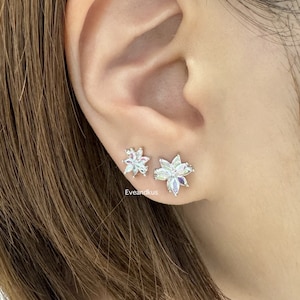 May include: A pair of silver flower-shaped earrings with clear cubic zirconia stones. The earrings are shown on a person's ear.