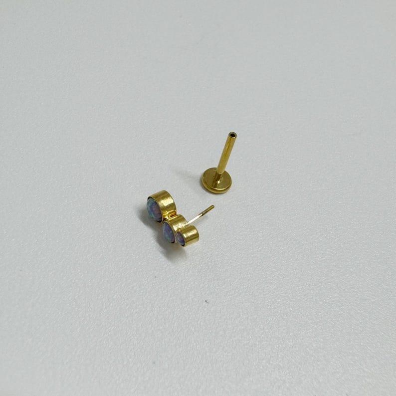 May include: A gold-toned body jewelry piece with three opal-like stones. The jewelry is separated into two parts: a post with a small ball on the end and a cluster of three stones.