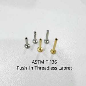 Könnte beinhalten: Fünf Labret-Stecker aus Chirurgenstahl und Gold, auch bekannt als Push-In-Threadless-Labrets. Die Stecker sind in einer Reihe angeordnet, wobei die goldenen Stecker an den Enden und die Stahlstecker in der Mitte liegen. Der Text "ASTM F-136 Push-In Threadless Labret" befindet sich unter den Steckern.