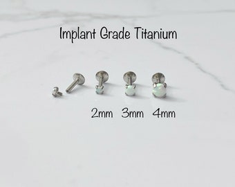 Puede incluir: Una colección de pendientes de titanio de grado implante con piedras de aspecto ópalo. Los pendientes se muestran en varios tamaños, incluyendo 2 mm, 3 mm y 4 mm. El texto "Implant Grade Titanium" se muestra.