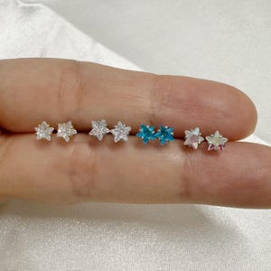 May include: A close-up of a hand wearing six pairs of star-shaped earrings. The earrings are silver, gold, and blue.