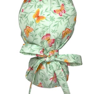 May include: A light green scrub cap with a repeating pattern of orange and pink butterflies and green foliage. The cap has a bow in the back for adjusting the fit. The fabric is a soft cotton.