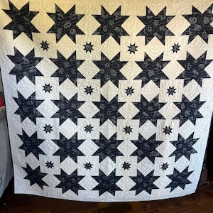 Puede incluir: Un edredón grande, hecho a mano, con un patrón repetitivo de estrellas azul marino sobre un fondo crema. Las estrellas están rellenas con un diseño blanco e intrincado. Estrellas más pequeñas se intercalan, creando un diseño simétrico y equilibrado.