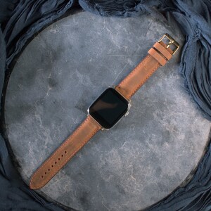 May include: A brown leather watch band with a gold-colored buckle is attached to a silver smartwatch. The watch and band are displayed on a gray surface, with a dark blue fabric draped around the edges.