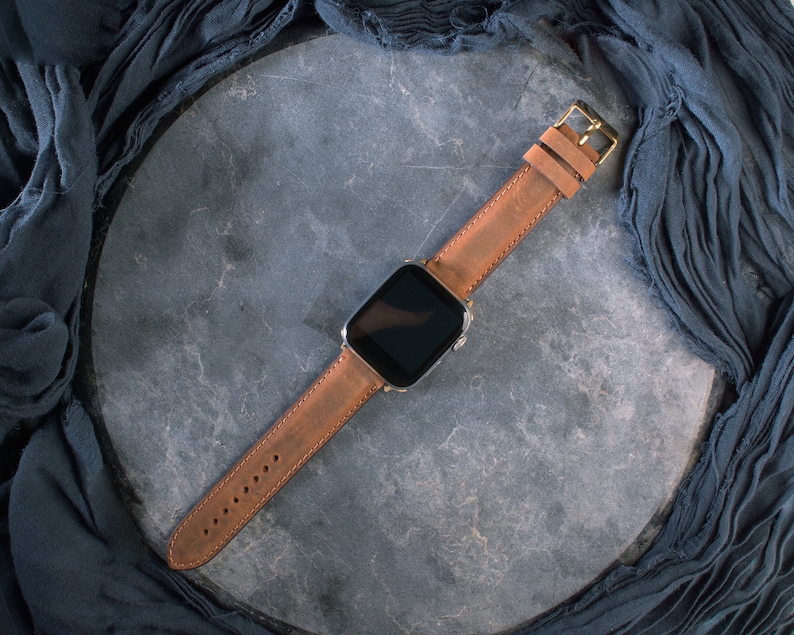 May include: A brown leather watch band with a gold-colored buckle is attached to a silver smartwatch. The watch and band are displayed on a gray surface, with a dark blue fabric draped around the edges.