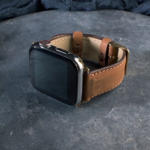 May include: A brown leather watch band with a stainless steel watch face. The band has a buckle closure and is made of a textured, distressed leather. The watch face has a black screen and a red button.