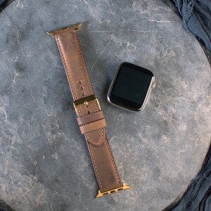May include: A brown leather watch strap with gold hardware for an Apple Watch.