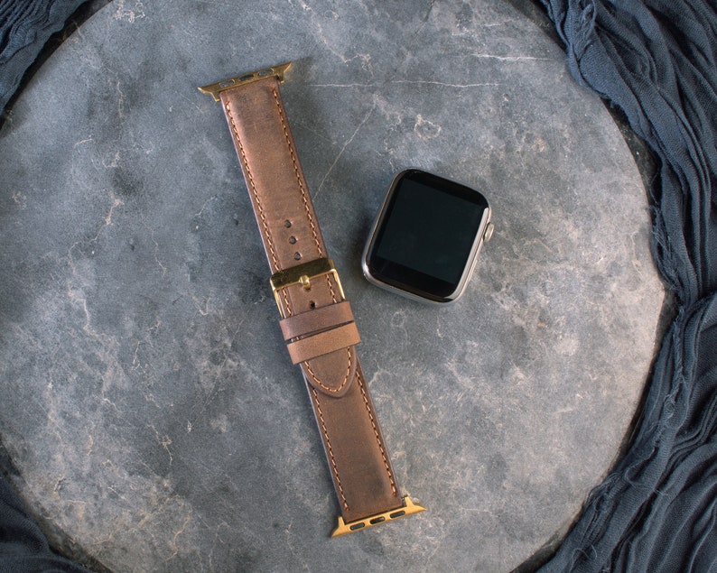 May include: A brown leather watch strap with gold hardware for an Apple Watch.