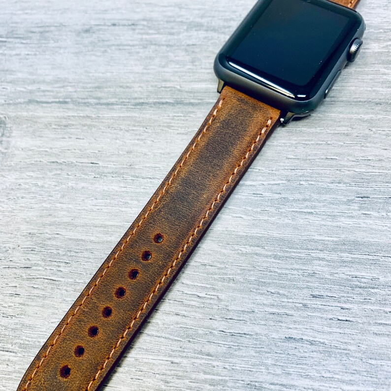 May include: A brown leather watch band with distressed detailing and stitching. The band is designed for an Apple Watch and has multiple holes for adjusting the fit.