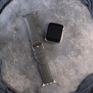 K&ouml;nnte beinhalten: Ein graues Lederarmband mit einer silbernen Schlie&szlig;e und einem schwarzen Zifferblatt.