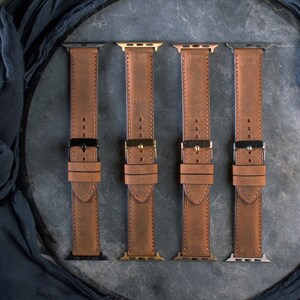 May include: Four brown leather watch bands with different metal hardware. The bands are made of distressed leather and have black, gold, silver, and gunmetal clasps. The bands are laid out on a dark gray surface.
