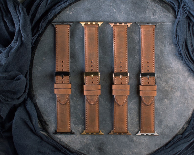 May include: Four brown leather watch bands with different metal hardware. The bands are made of distressed leather and have black, gold, silver, and gunmetal clasps. The bands are laid out on a dark gray surface.