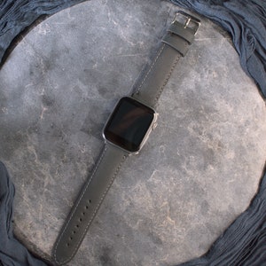 K&ouml;nnte beinhalten: Ein schwarzes Lederarmband mit einem silbernen Zifferblatt. Die Uhr liegt auf einem grauen Hintergrund.