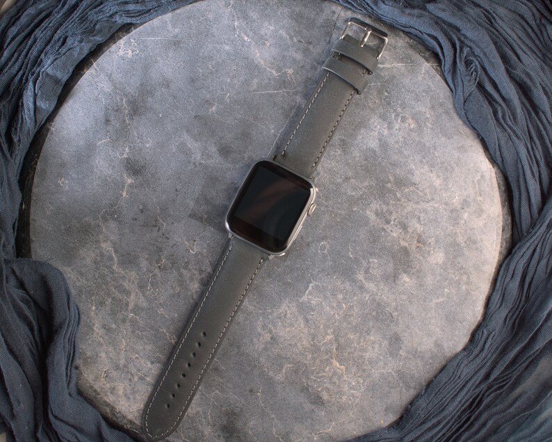 K&ouml;nnte beinhalten: Ein schwarzes Lederarmband mit einem silbernen Zifferblatt. Die Uhr liegt auf einem grauen Hintergrund.