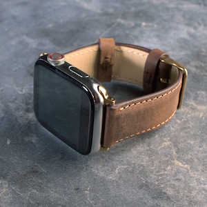 May include: A brown leather watch band with a silver Apple Watch face. The band has a stitched edge and a gold buckle.