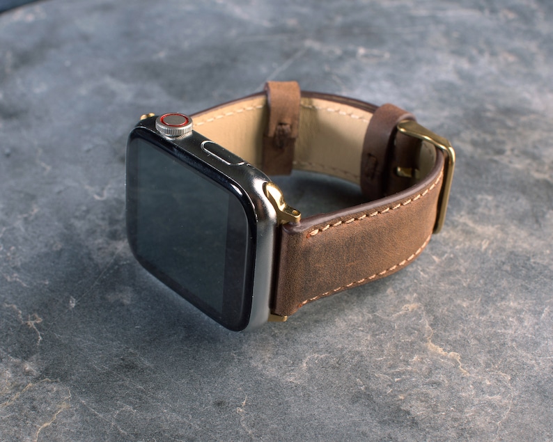 May include: A brown leather watch strap with a silver Apple Watch face. The strap has a stitched edge and a gold buckle.